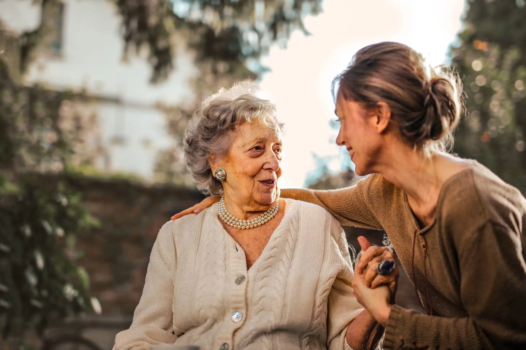 An adult daughter hugging her older mother in a sunny garden during a visit.